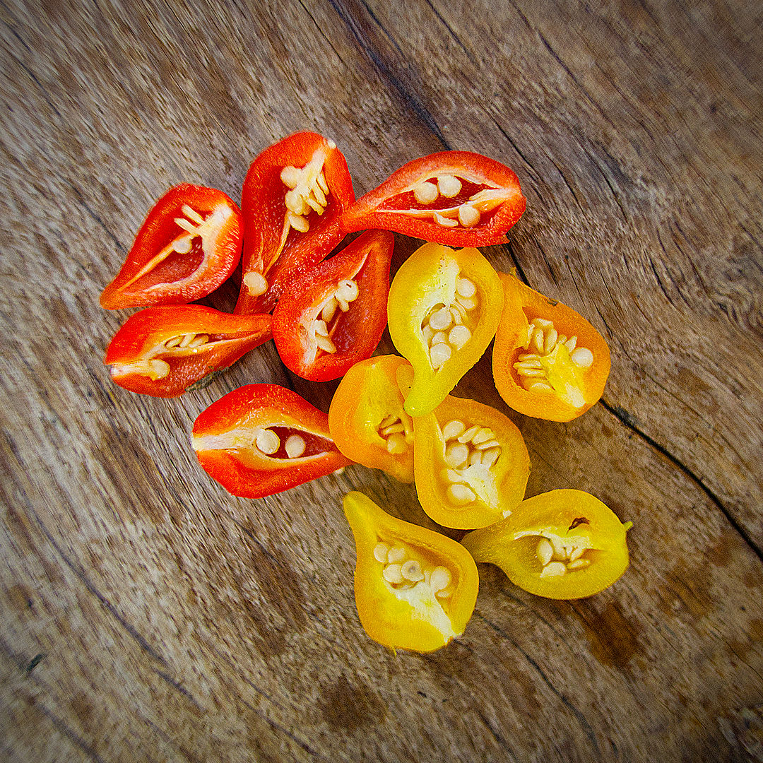 World of Chillies: Red and yellow Biquinho chillies sliced in half on a wooden board, showing their tiny seeds. 